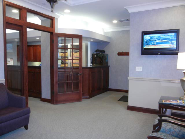 The image shows an interior space that appears to be a waiting room or lobby within a building, featuring a large wooden door with glass panels, a comfortable seating area with a couch and armchairs, a television mounted on the wall, and a counter or desk with a monitor displaying what seems to be a video or presentation. The color scheme includes neutral tones with wood elements and is well-lit.