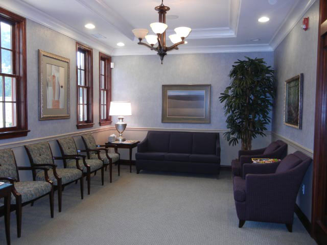 The image shows an interior view of a waiting room with a couch, chairs, a coffee table, a lamp, a framed picture on the wall, and a large window allowing natural light into the space.