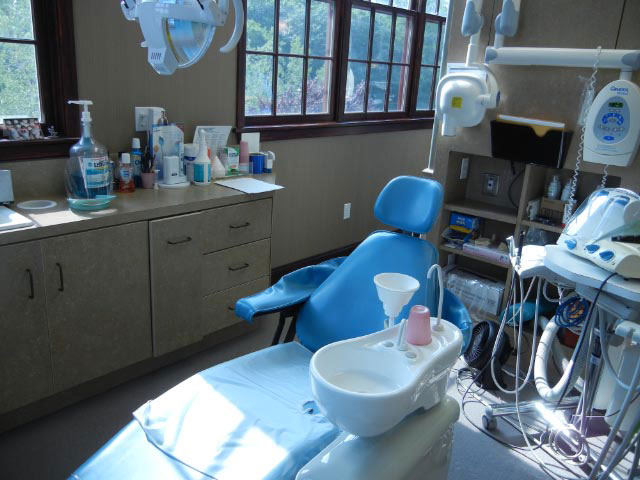 The image shows a dental office interior with a large blue chair in the foreground, an open door leading to a window view, and various medical equipment and supplies surrounding the chair.