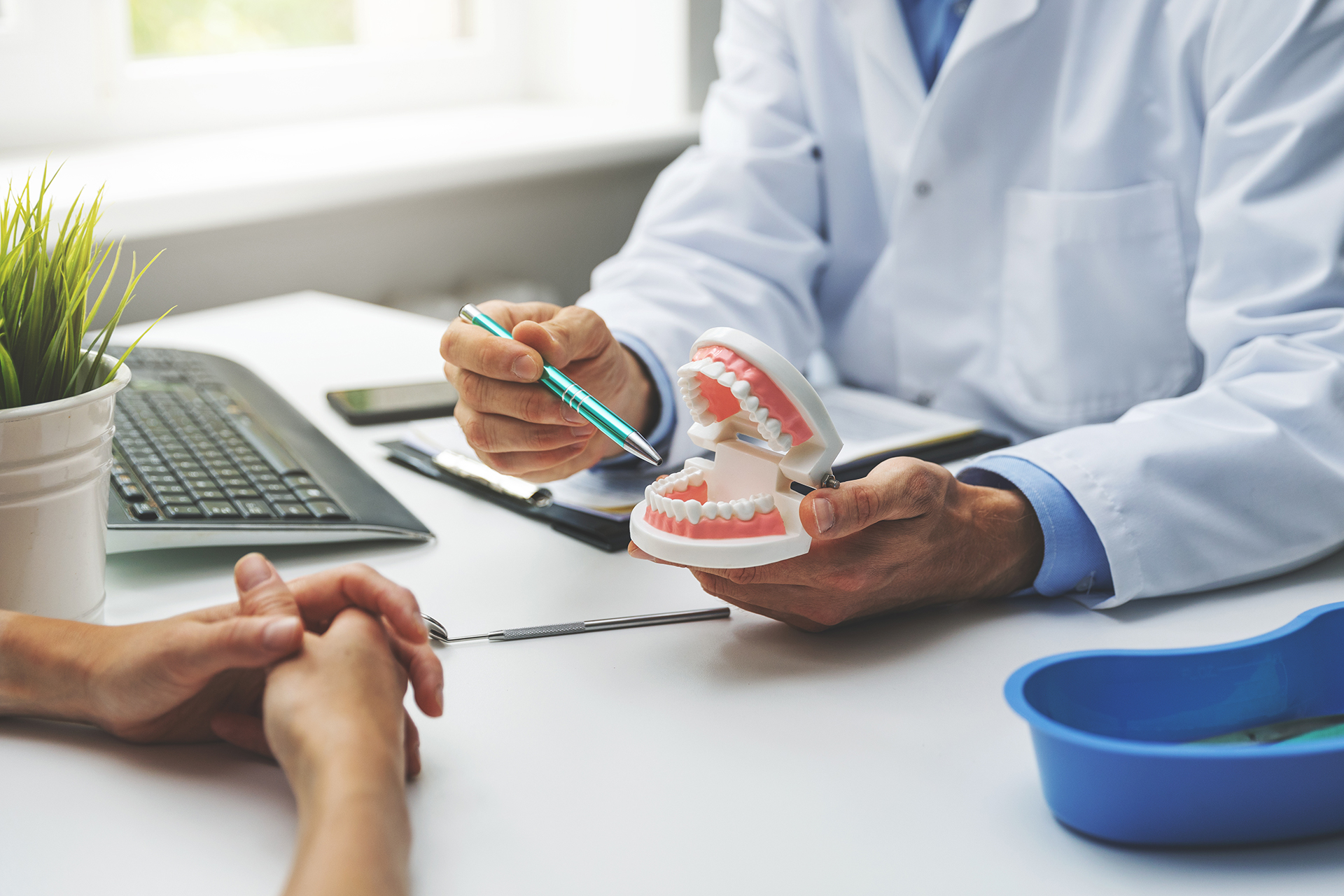 A dental professional examines a patient s teeth while seated at a desk with a laptop and office supplies.