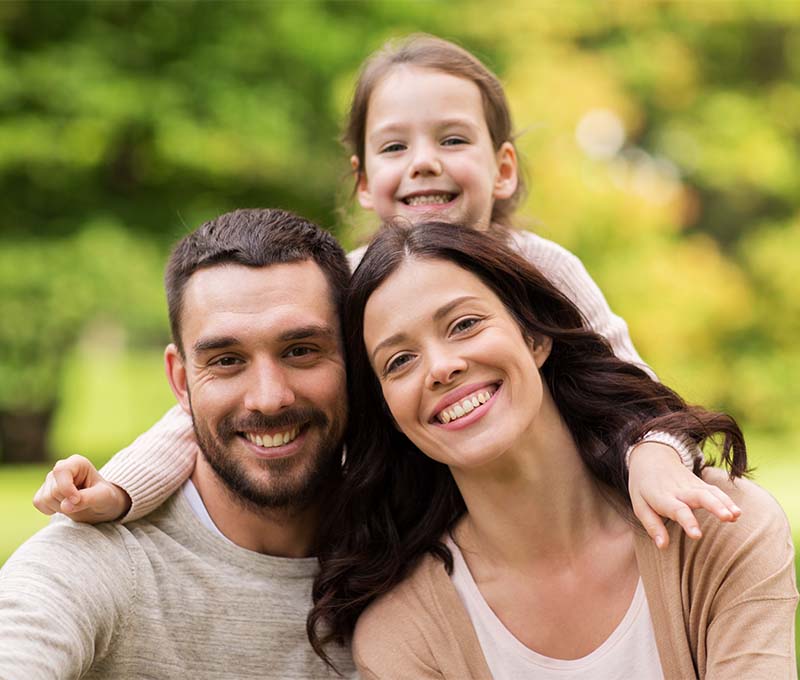 The image depicts a family of four with two adults and two children, smiling and posing together in an outdoor setting during daylight.