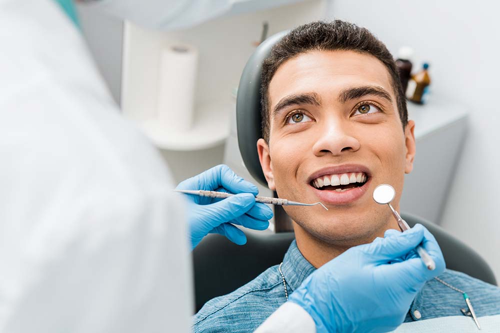 The image shows a person sitting in a dental chair with their mouth open while receiving dental treatment from a dentist wearing protective gloves, who is standing behind them.