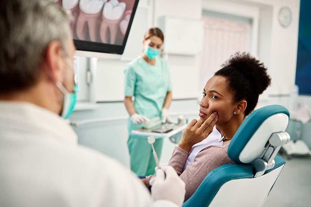 The image shows a dental office setting where a patient is seated in a chair with a dental professional standing nearby, both wearing surgical masks and face shields.
