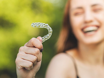 A smiling woman holding up a clear plastic retainer with her right hand against a blurred outdoor background.