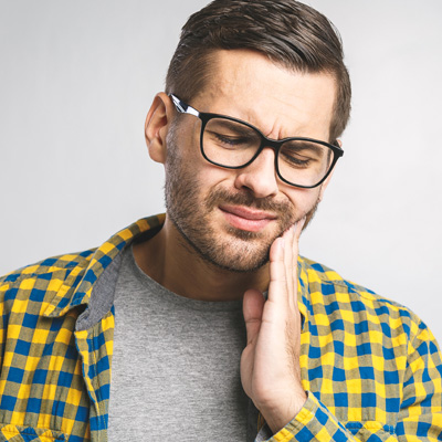 The image shows a man with glasses and a beard, wearing a plaid shirt, holding his hand to his mouth, appearing to be in pain or discomfort, likely due to dental issues.